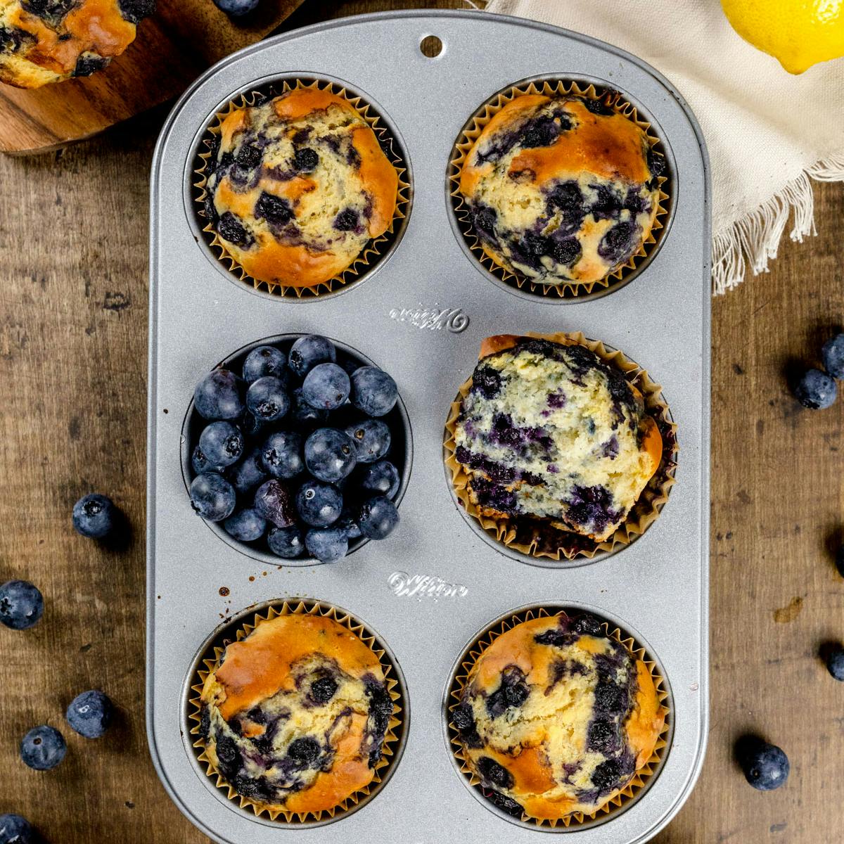Six blueberry protein muffins in a muffin tray with fresh blueberries. 