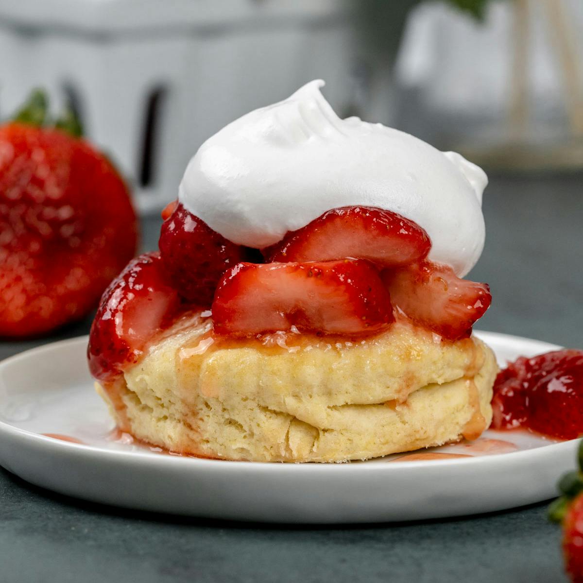 A vegan strawberry shortcake biscuit is on a white plate covered with strawberries and dairy free whipped cream. Strawberries are blurred in the background. 