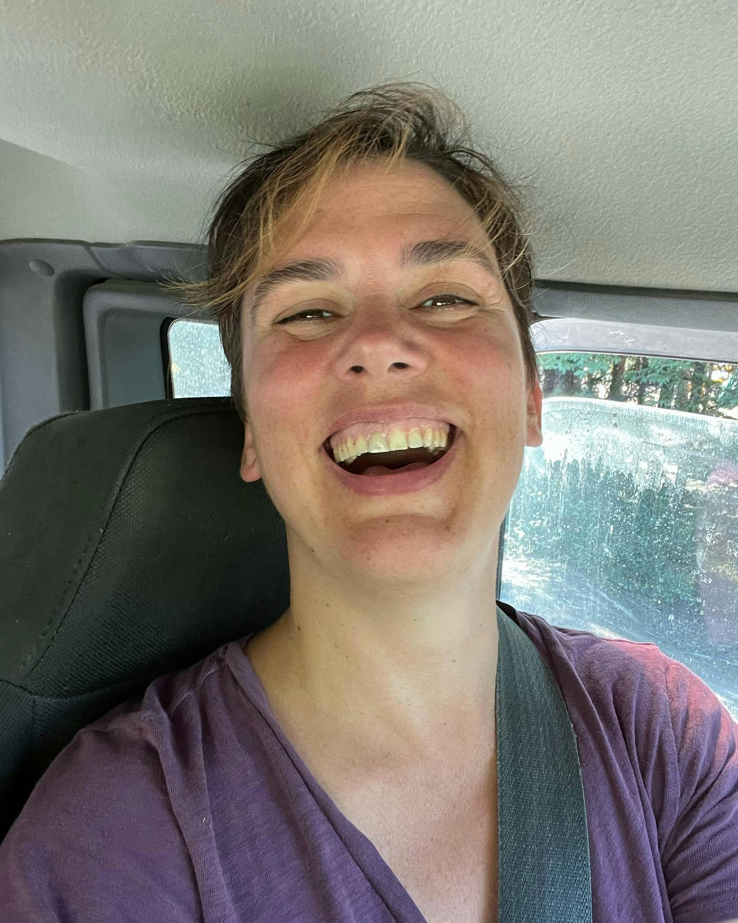 Janelle Hardy, a white woman with short brown hair and a summertime tan, is sitting in a truck with muddy windows, wearing a purple t-shirt, with her head tossed back and a huge, eye-crinkling smile on her face.