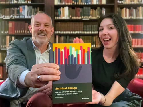 Josh Clark and Veronika Kindred hold a copy of Sentient Design in front of a vast bookshelf