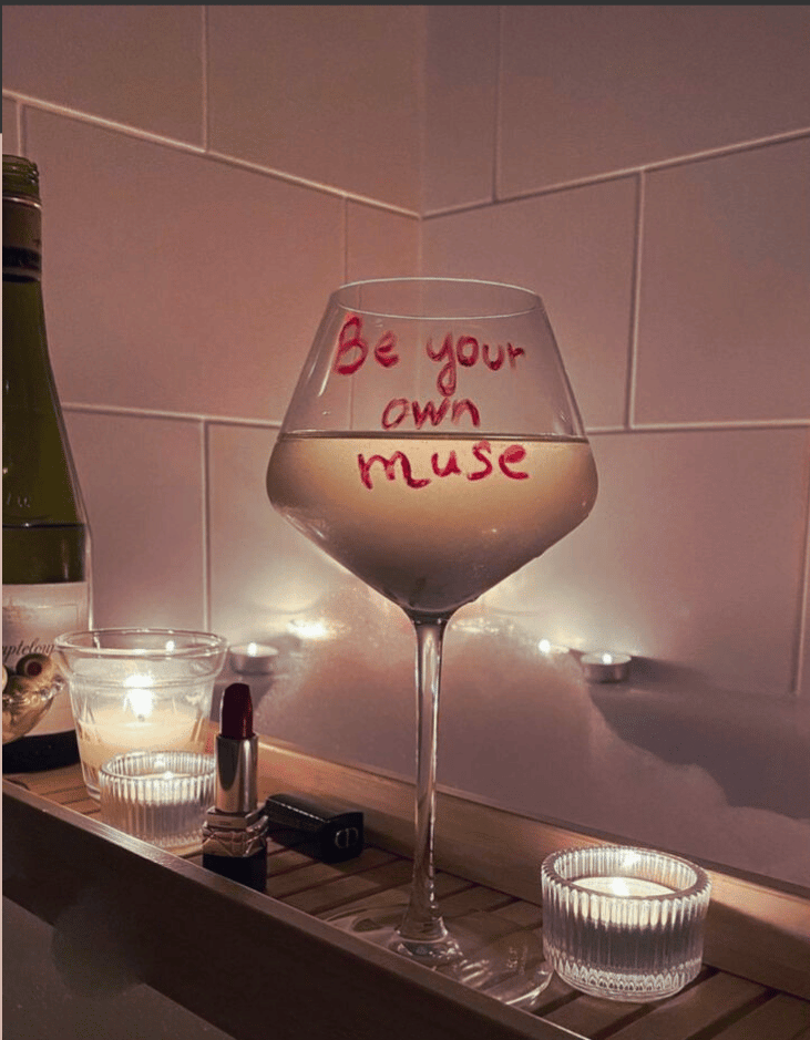 A wine glass filled with liquid has the words "Be your own muse" written on it. It is placed on a wooden tray alongside a bottle, two candles, and a tube of lipstick. The setting appears cozy, with soft lighting. Below the image, text reads: "Self-Worship Journal for High-Achieving Women. A powerful journaling guide to help you rebuild self-trust, embody confidence, and step into your most magnetic, untouchable self. Michelle Burke." The background and border consist of muted, earthy tones.