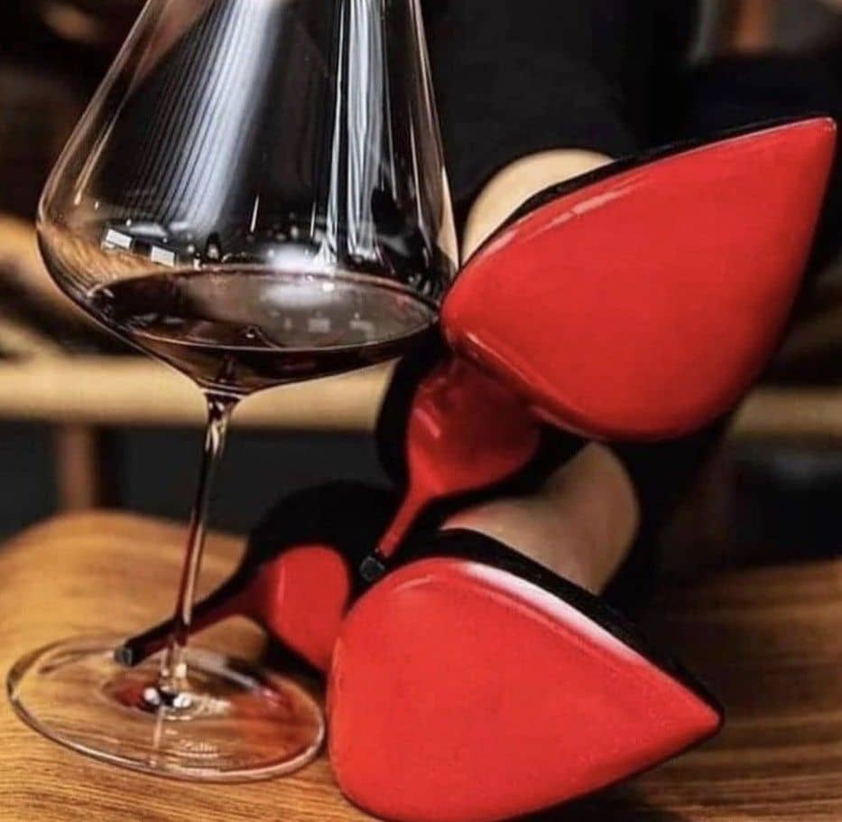 A glass of red wine is placed on a wooden surface next to a pair of black high-heeled shoes with distinctive red soles. The focus is on the contrasting colors and elegant composition.