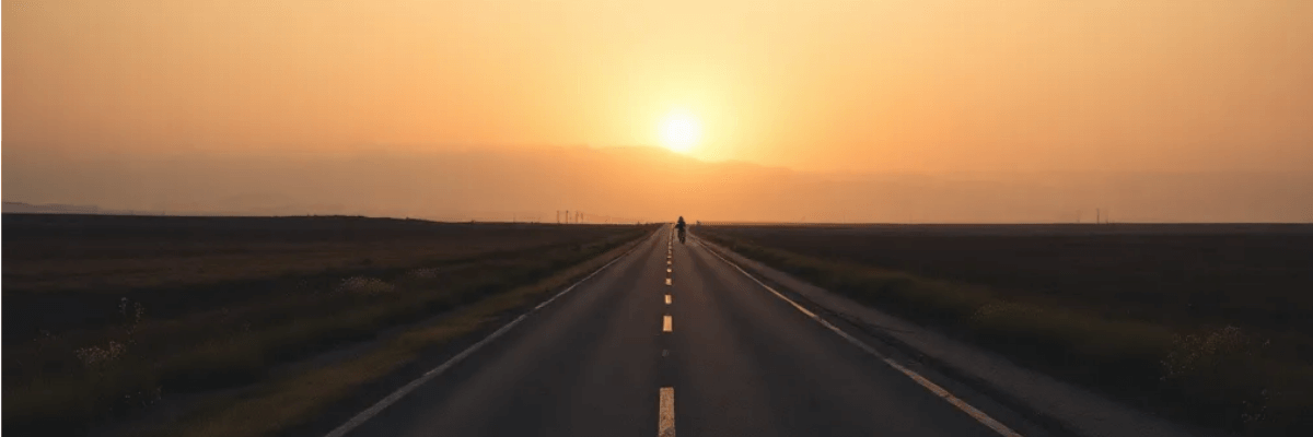 A long, straight road stretches toward the horizon as the sun rises, casting warm light over distant, misty mountains and open fields on either side.