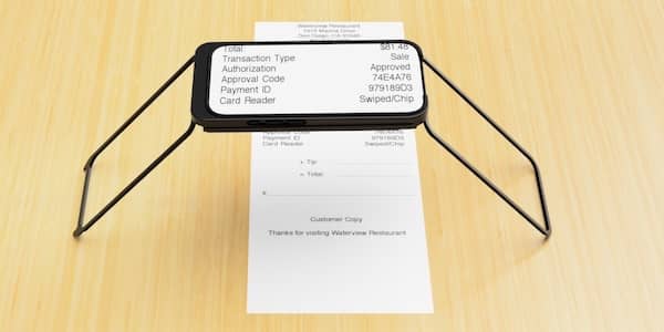 An iPhone is mounted in a black Case for Vision stand with its articulated legs extended on a tabletop. Beneath the phone is a printed receipt with small text. The phone’s screen displays a magnified, high-contrast view of the receipt, clearly showing details like the total, authorization code, and payment type. The setup allows for hands-free magnification of printed materials.