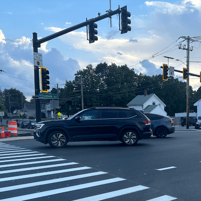 Cars zoom through a busy intersection with a brightly painted crosswalk and overhanging set of stoplights