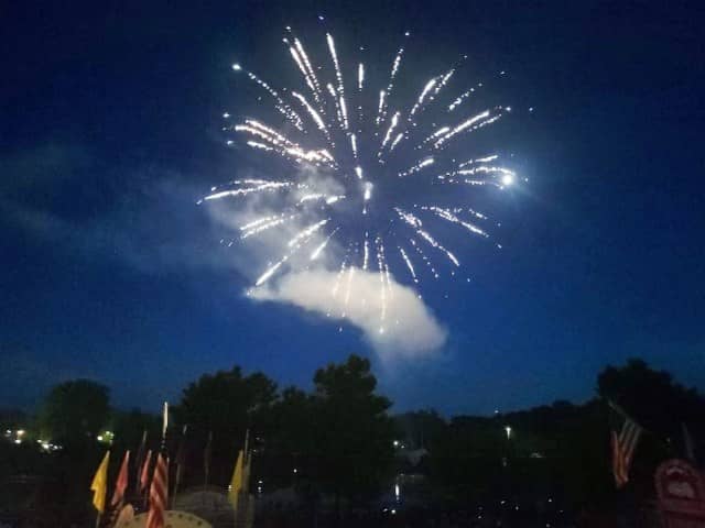  A bright white firework explodes into a dark blue night sky, sending streaks in every direction. Below, several people look up at the site against a silhouetted backdrop of trees.
