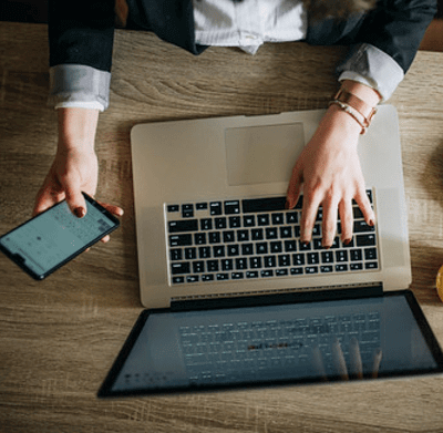 Looking down on a woman's hands holding a smartphone and typing on a laptop on a wooden desk
