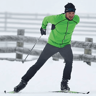 A man skate skiing through falling snow in front of a fence pushes of his right leg and ski to propel himself forward