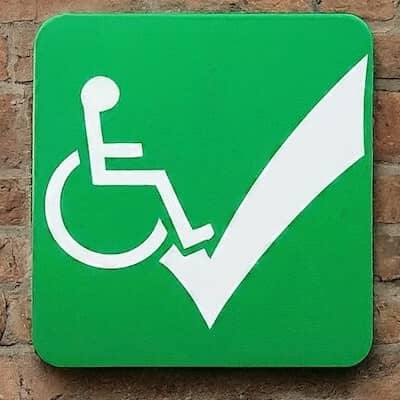 A green sign with the universal symbol of accessibility of a person in a wheelchair and a big checkmark hangs on a brick wall