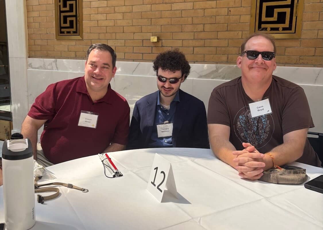 Simon, Zach, and Garin smile at a round table with a "12" place card, name badges visible, at the MCB summer internship closing ceremony under a marble and brick colonnade.