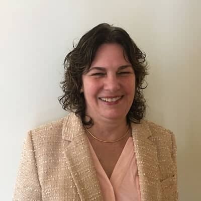 Headshot of DeAnn Elliott, a woman with wavy brown hair, smiling and wearing a light pink blouse under a beige textured blazer. 