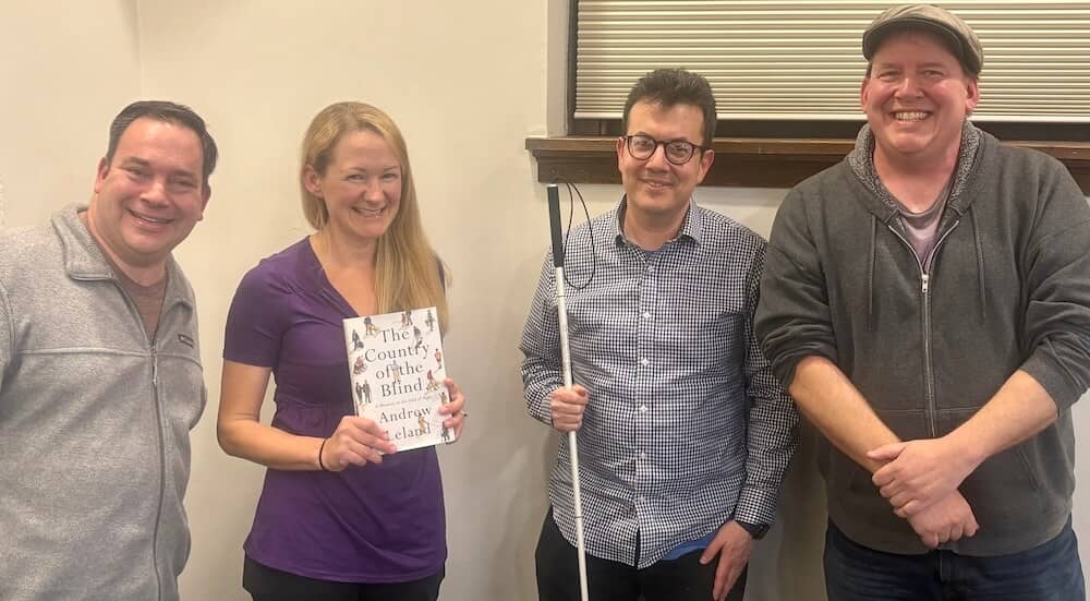 Four people smiling and standing together indoors after a talk. The second person from the left is holding a copy of the book The Country of the Blind by Andrew Leland, who stands next to her holding a white cane.