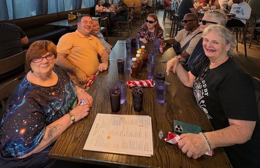 A group of eight friends seated at a long wooden restaurant table, smiling toward the camera. Purple and amber drinks, hot sauce bottles, and red-checked napkins line the table, with a menu resting in the middle. Other diners are visible in the background