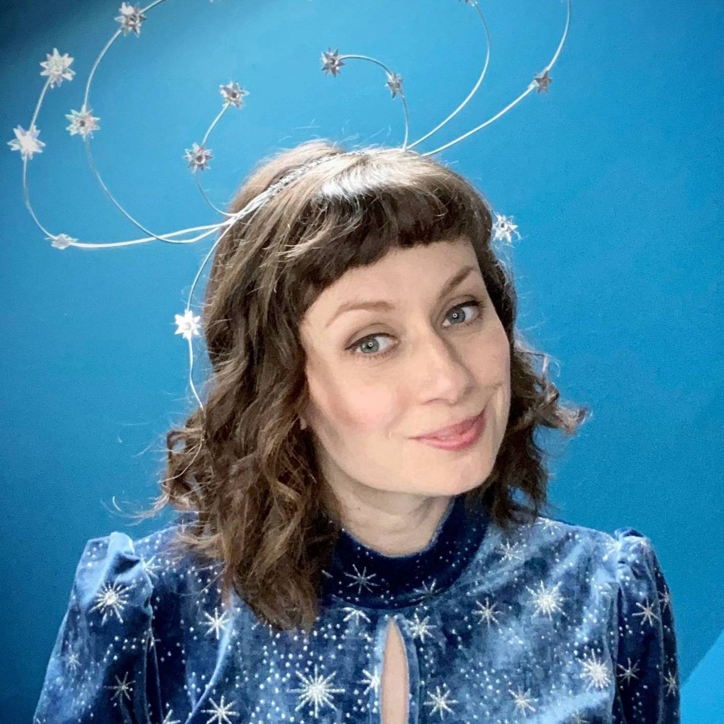 A person with wavy hair is wearing a celestial-themed headpiece featuring wire loops adorned with star shapes. They are dressed in a blue outfit with white star patterns and a keyhole neckline. The background is a gradient of blue shades.