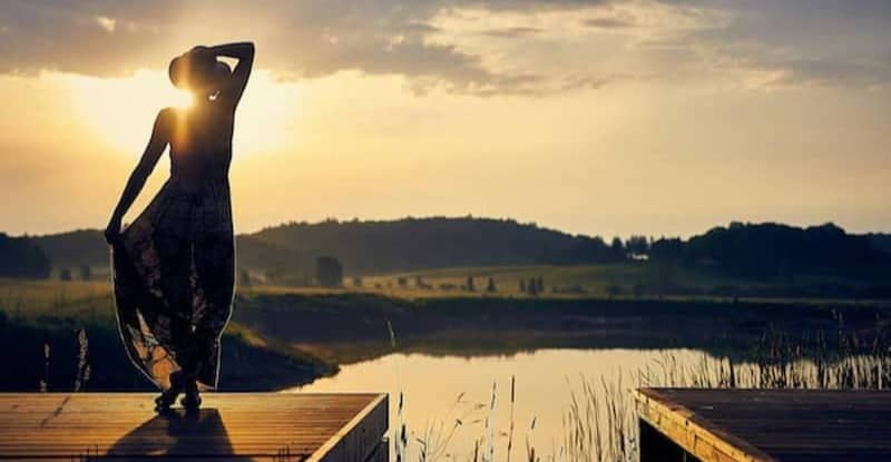 Woman standing in front of a lake while holding her hat with one hand and her dress with the other one
