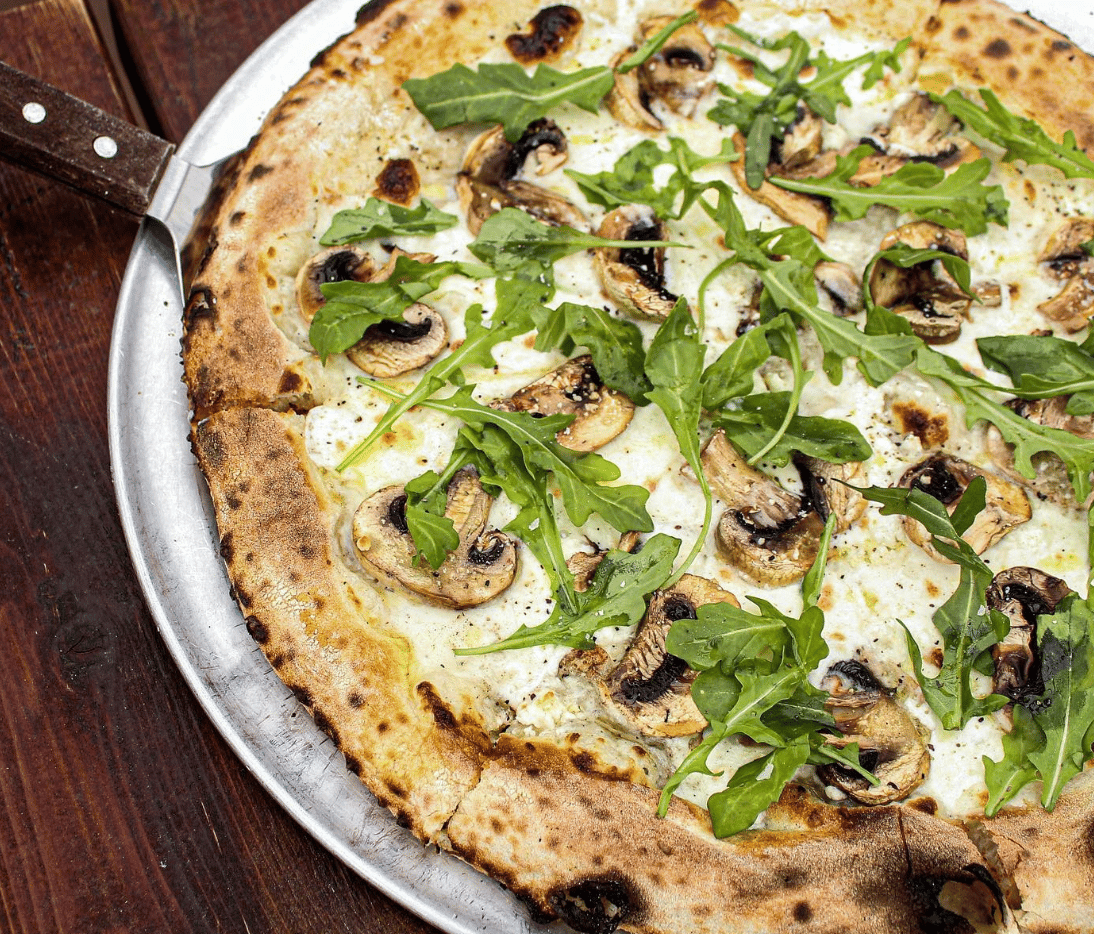 pizza fresh from the oven on a silver tray, covered in white sauce, mozzarella, arugula, and sliced mushrooms
