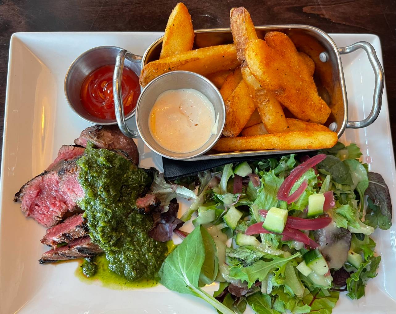 dinner plate with a sliced steak topped in green chimichurri with side of large cut fries and side salad