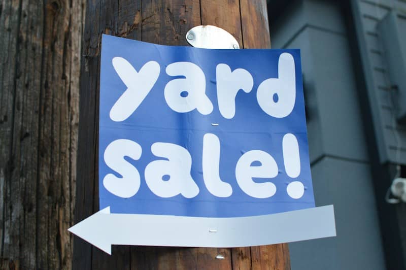 A blue yard sale sign with white text attached to a wooden pole