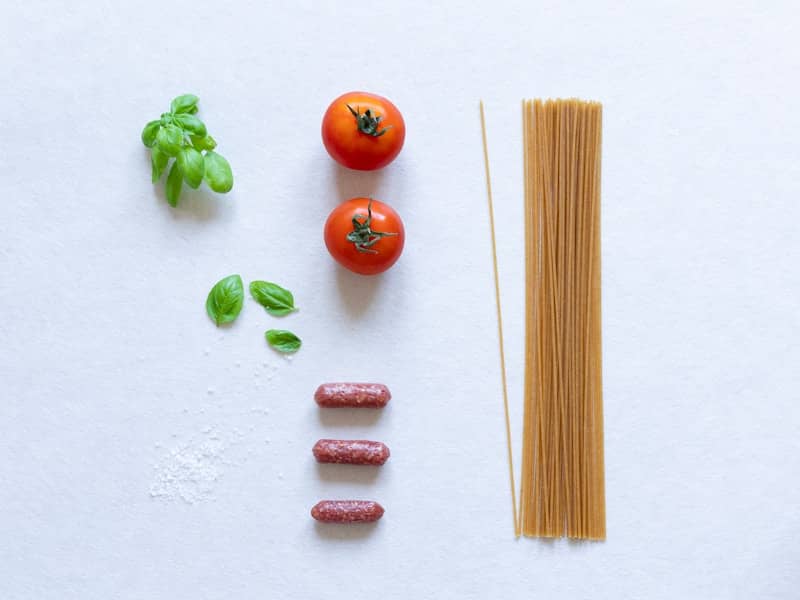 Tomatoes, basil, sausages, and spaghetti arranged on white.