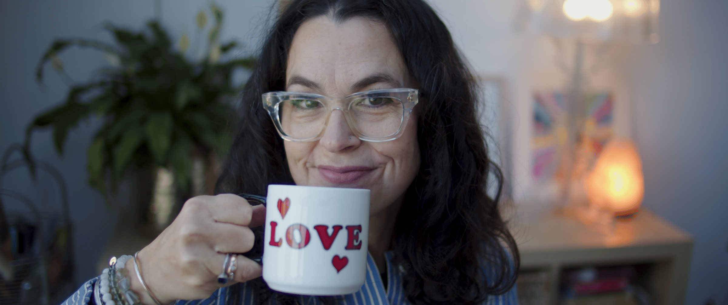 woman with dark hair and clear glasses smiling as she holds a coffee cup to her mouth.