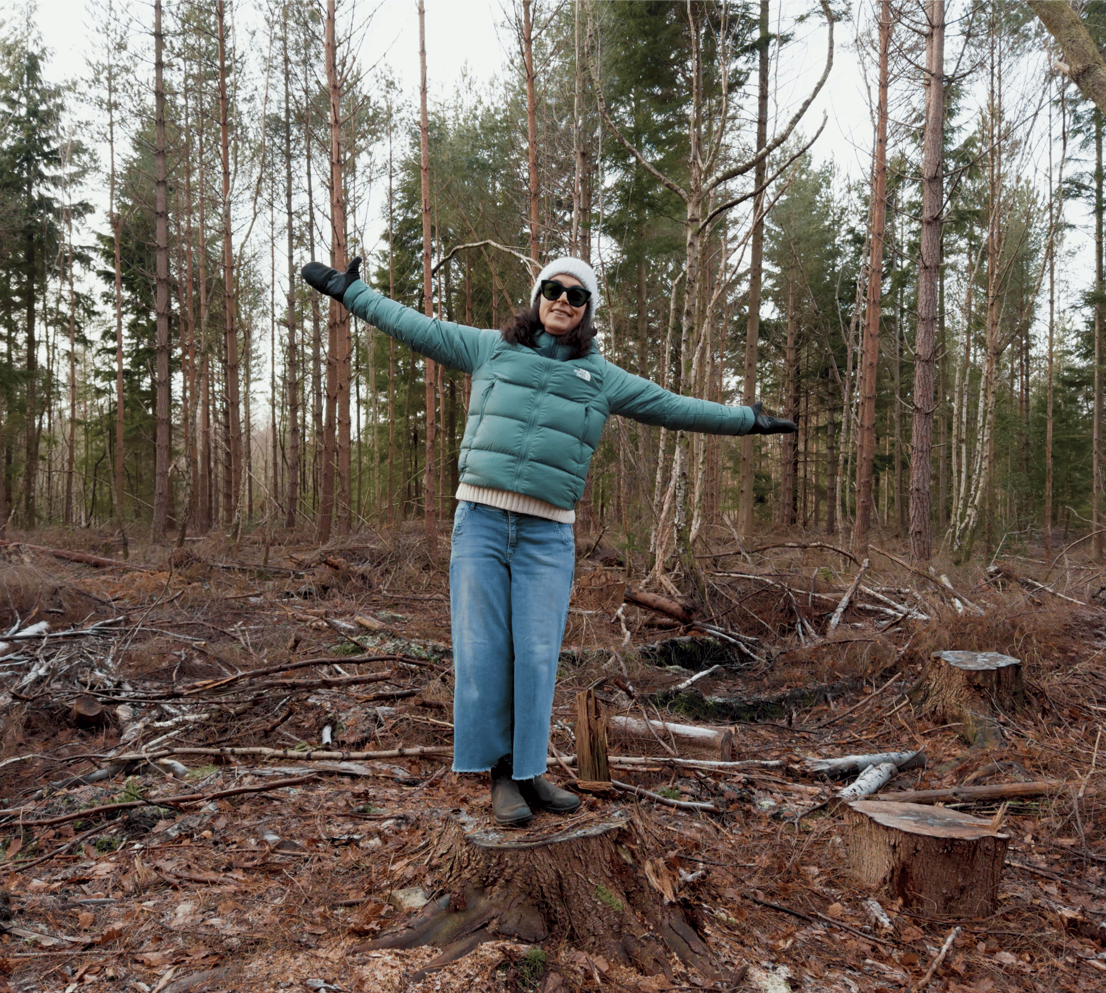 woman standing in the woods wearing green puffy jacket, a hat and gloves, standing on a treestump.