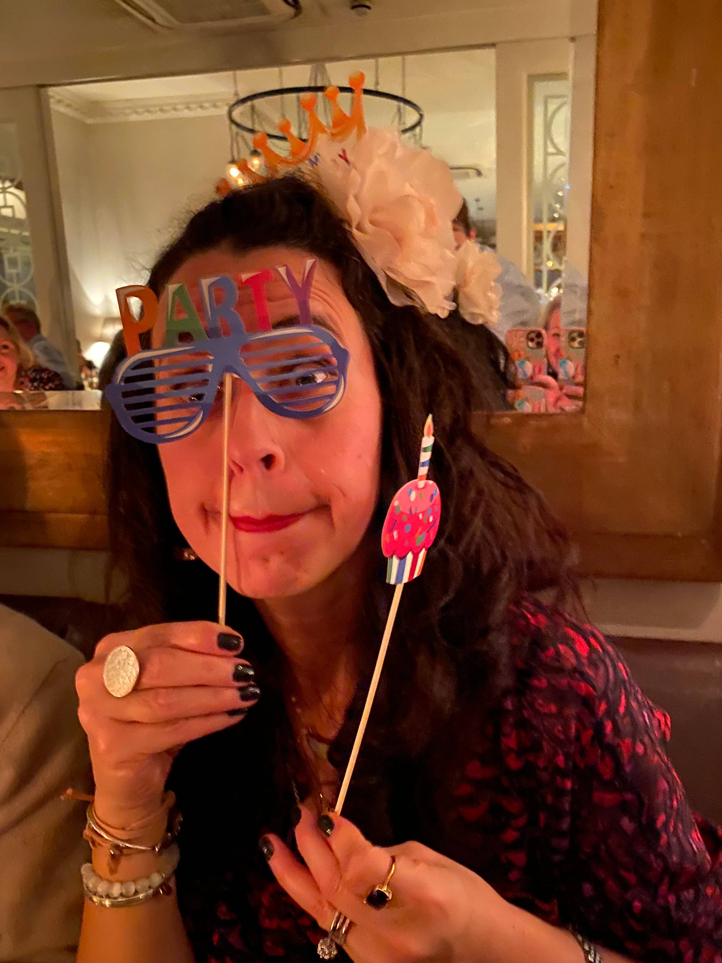 Woman with birthday glasses on holding a cupcake on a stick.