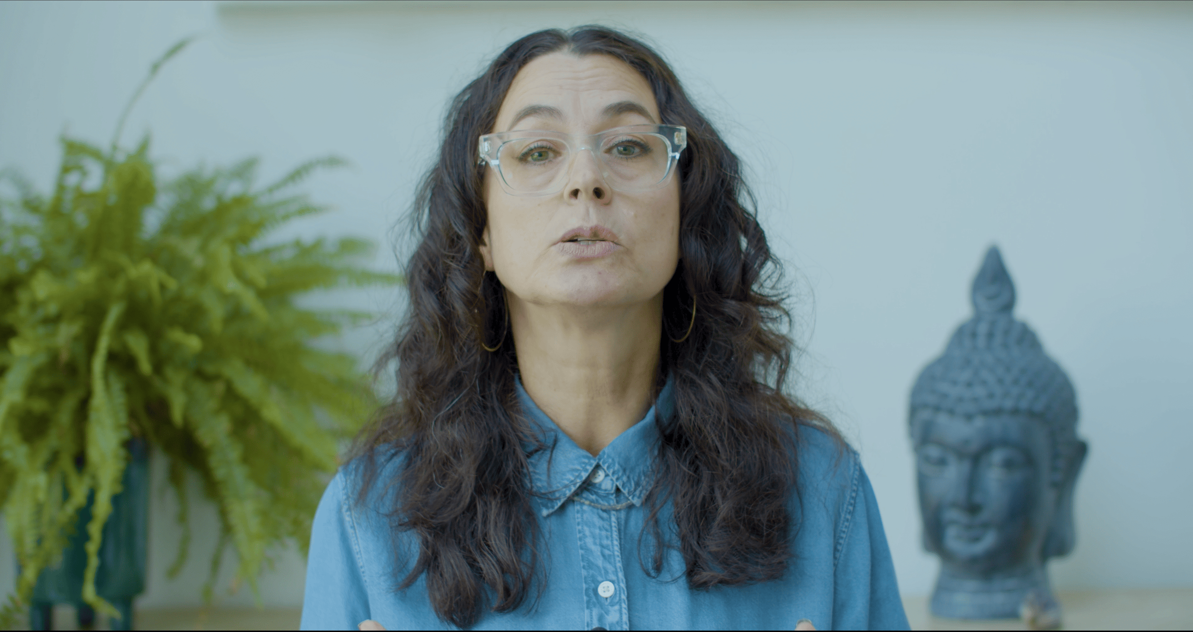 woman in a denim shirt with dark brown hair and clear glasses.