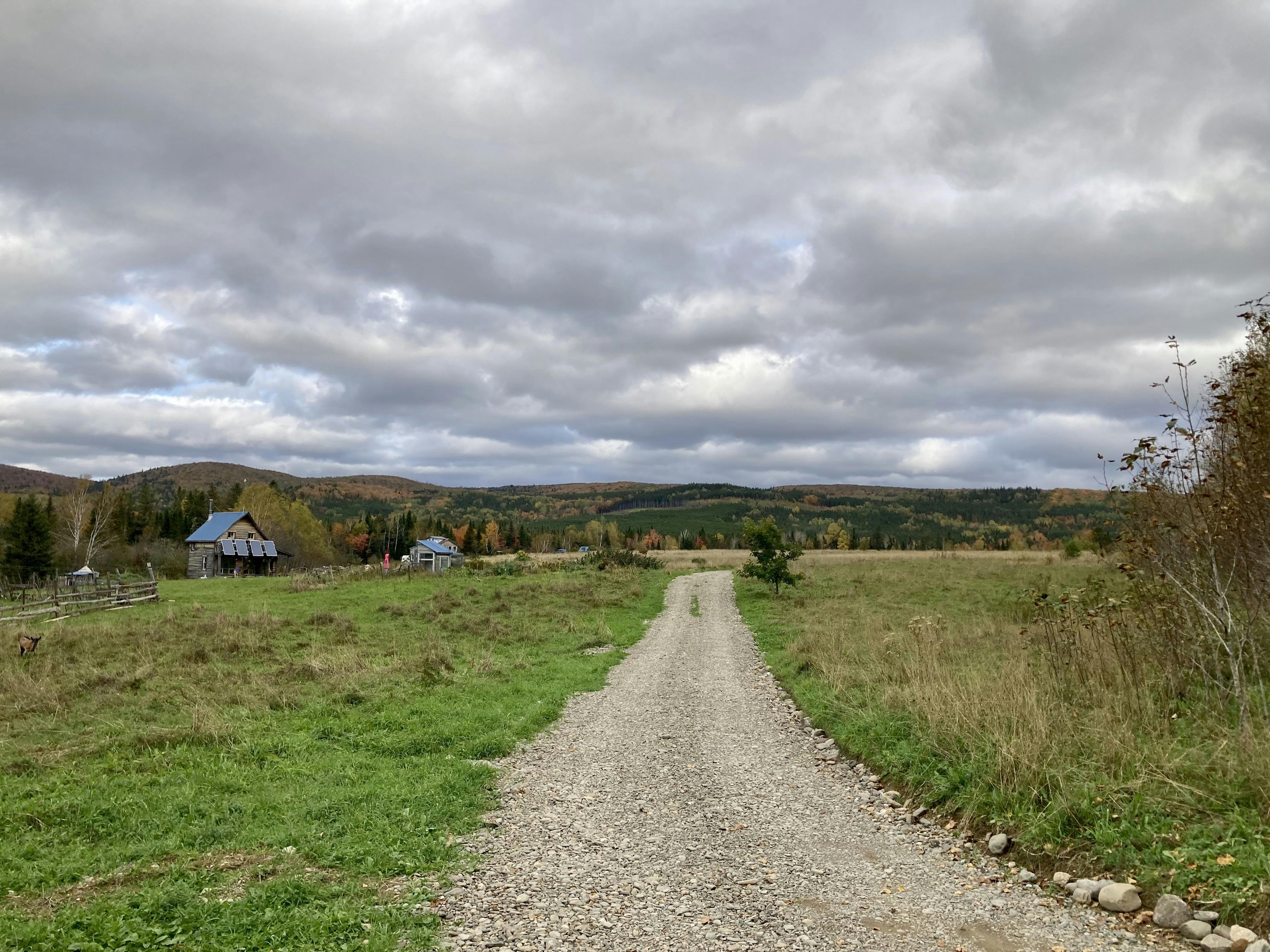 A photo of a dirt road going across a field with cabins on the left and hills in the distance.