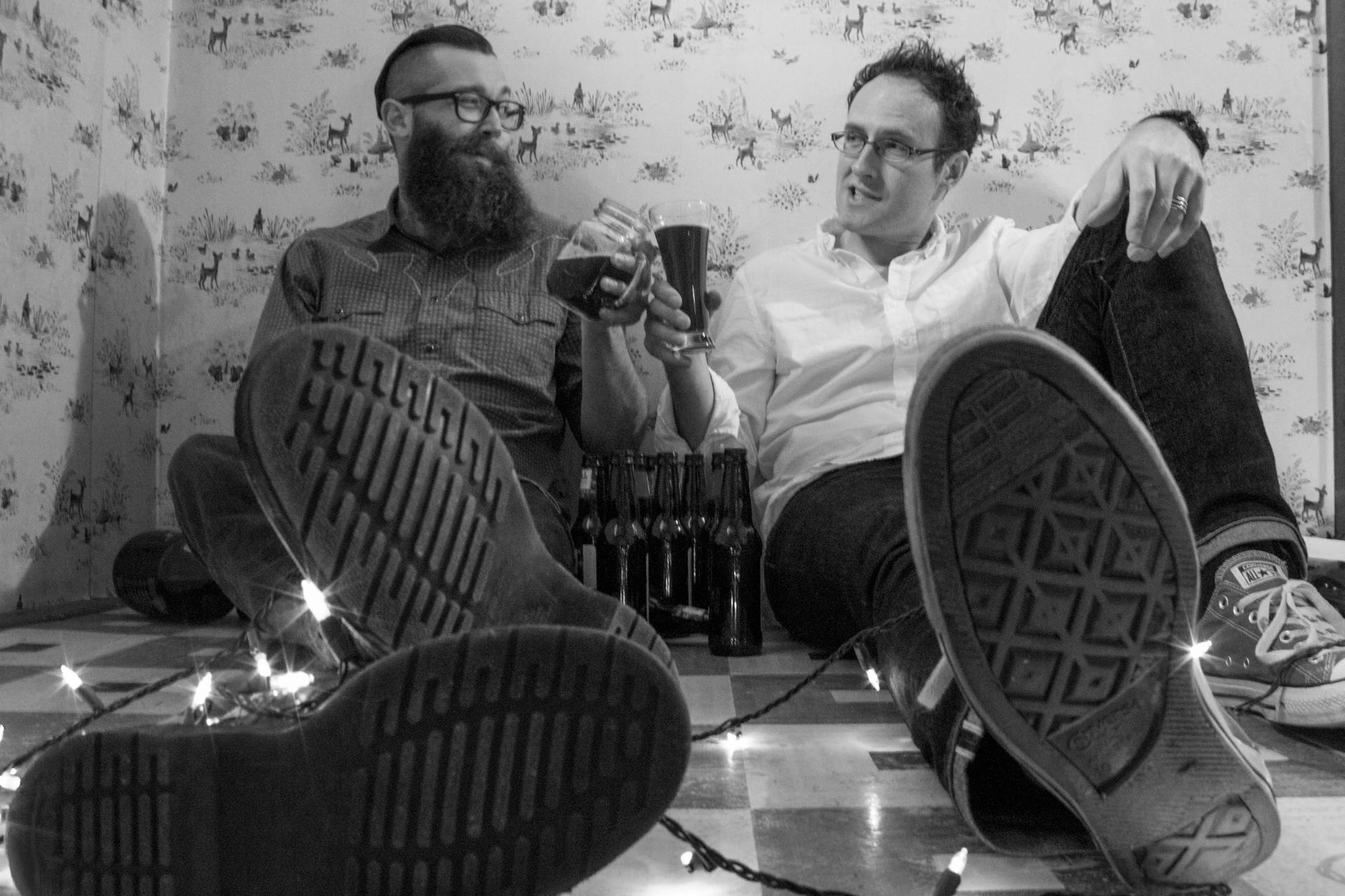 A picture of two boozy podcasters clinking glasses on the floor with some epic baby deer wallpaper in the background and christmas lights draped around their legs