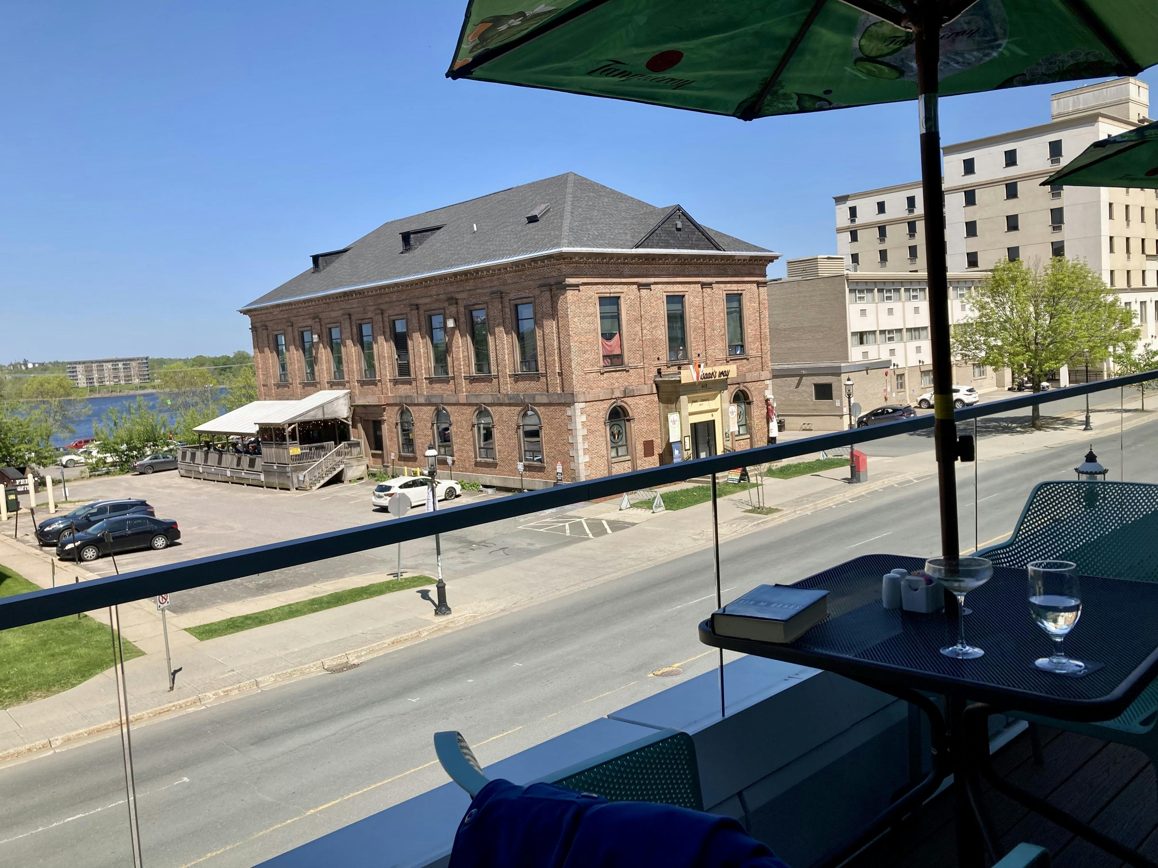 A photo from a second-storey patio, overlooking a street, a heritage building, a mid-century hotel, and the Wolastoq River