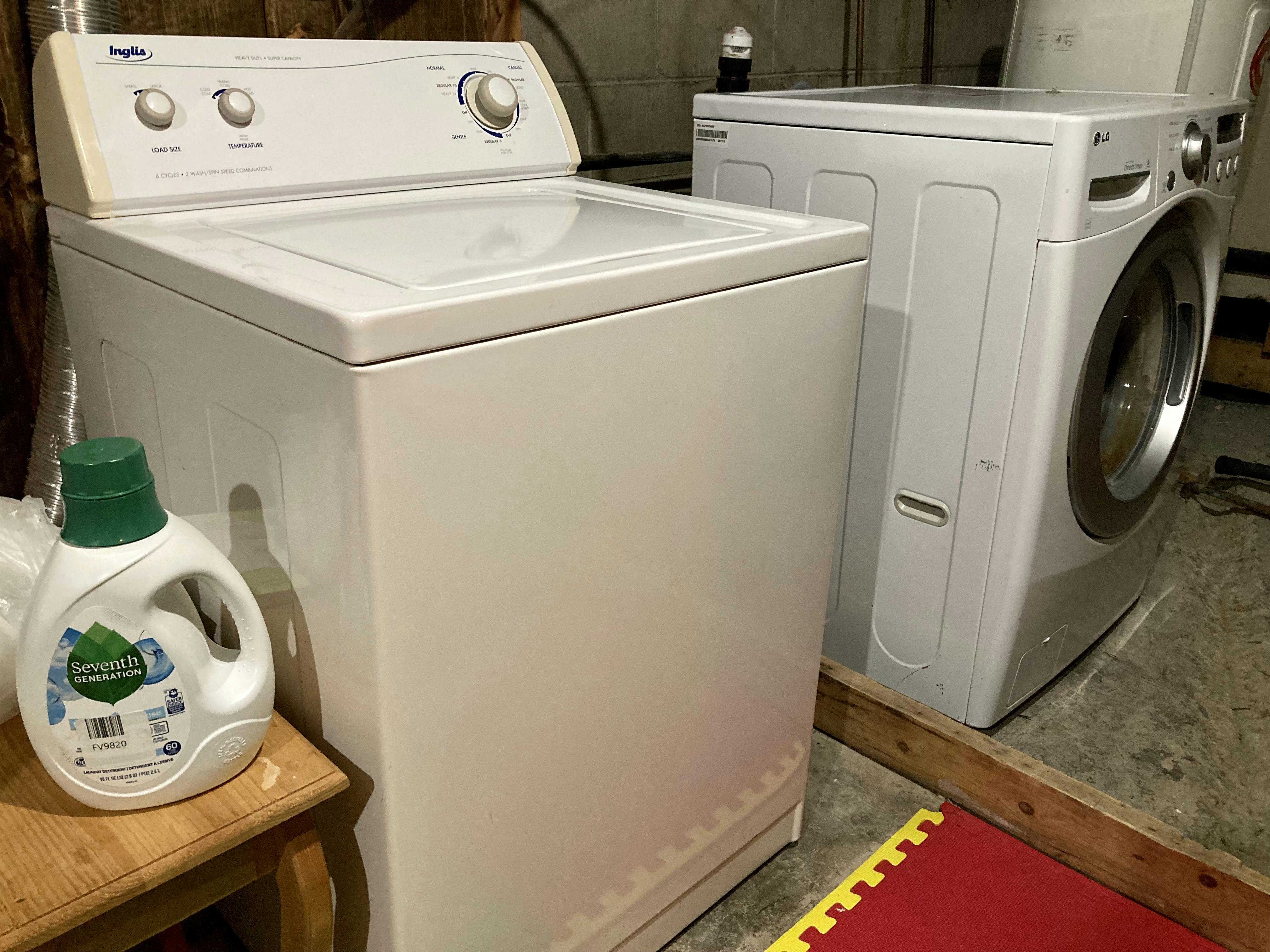 A photo of  an old washing machine in use while a newer model sits, discarded, behind it.