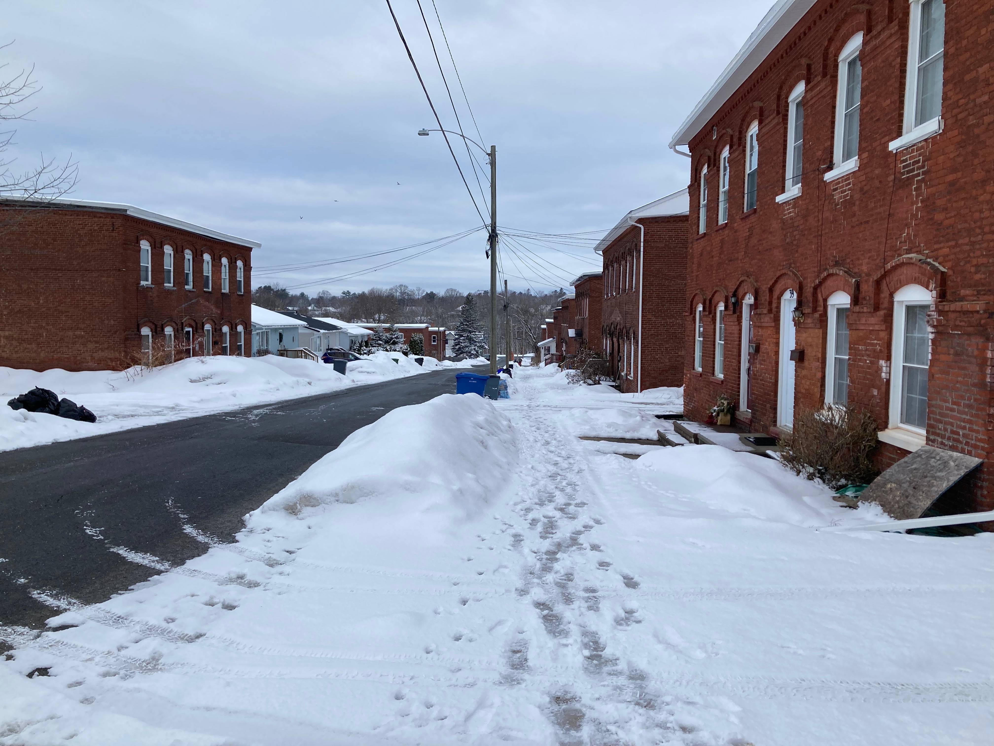 A photo of a view down a sidewalk with snow and footprints all over it and brick houses on either side of the street