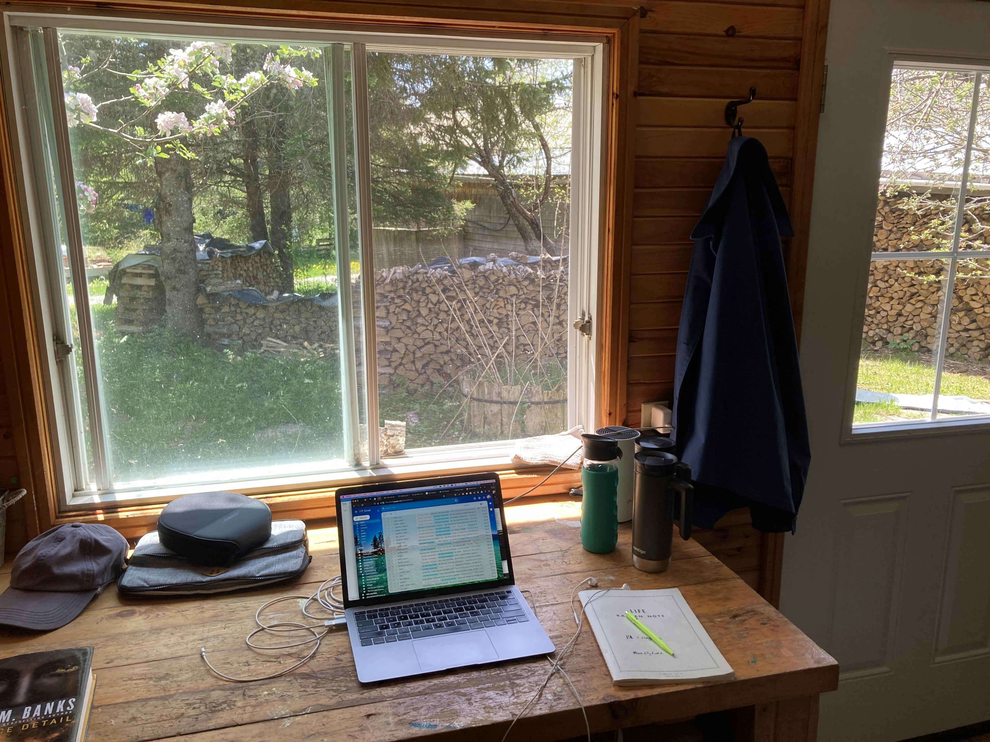 A photo of a rustic desk inside a rural cottage showing a computer, coffee and water containers, a notebook and pen, headphones, a hat, and a sci-fi book