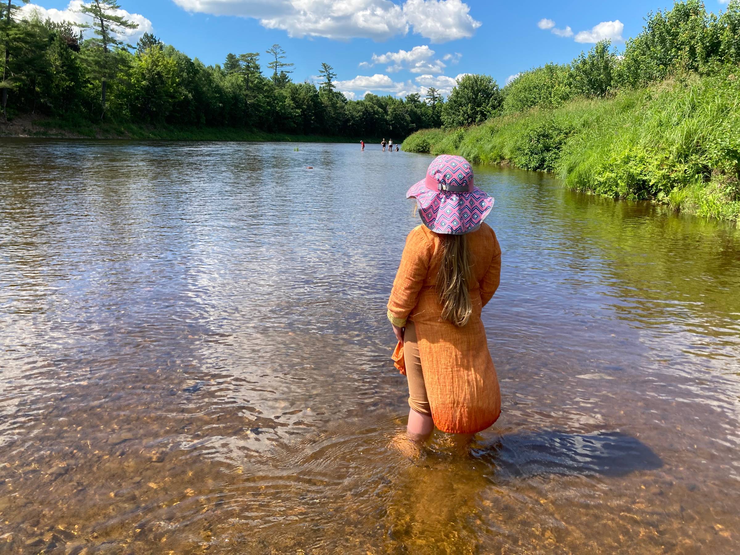 A young child with a ponytail and a pink hat and an orange dress stands in a river, looking away from the camera at a group of people upstream under a blue sky with white fluffy clouds.