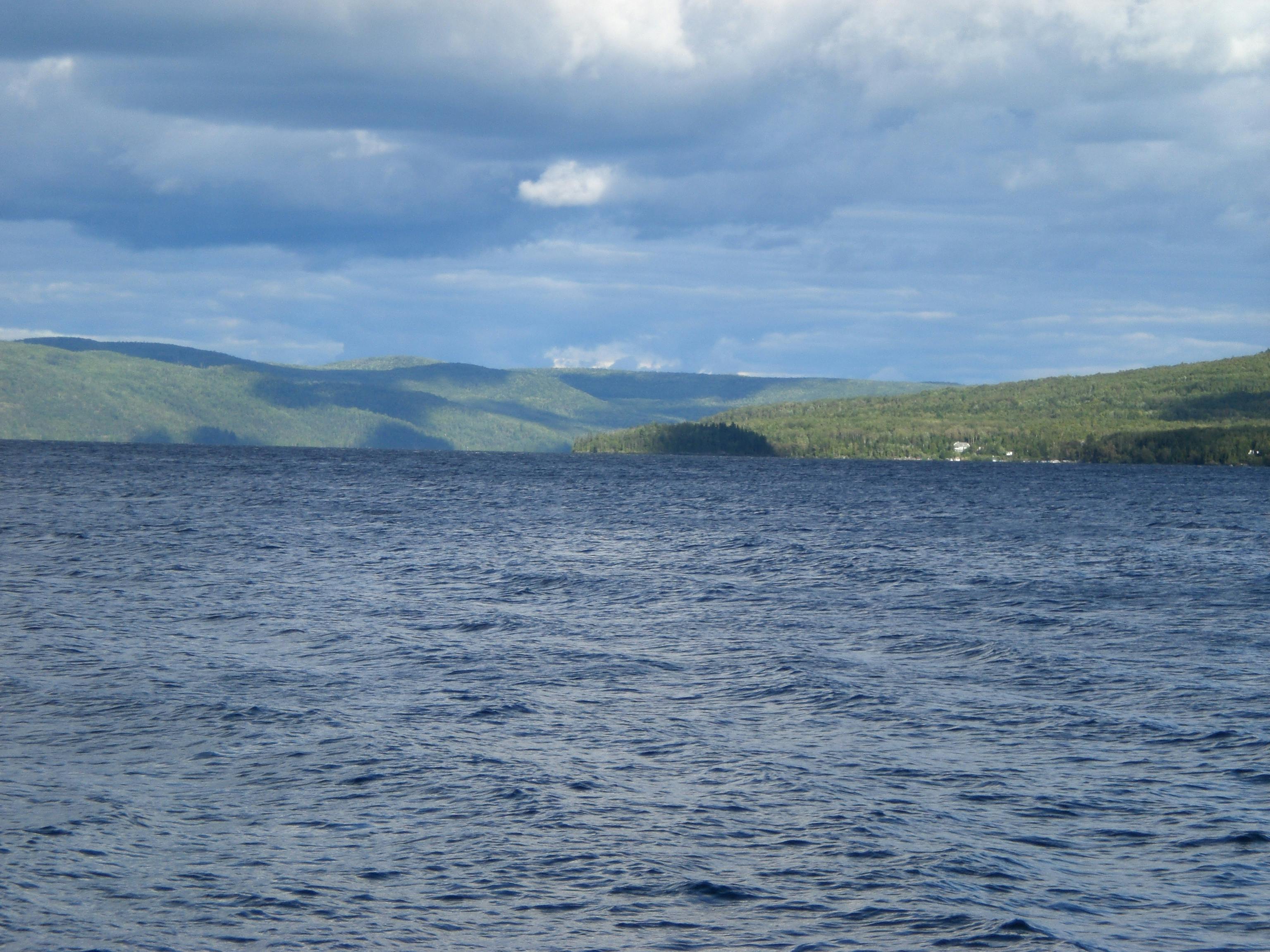 A photo of a lake with green hills in the background, and grey clouds overhead.