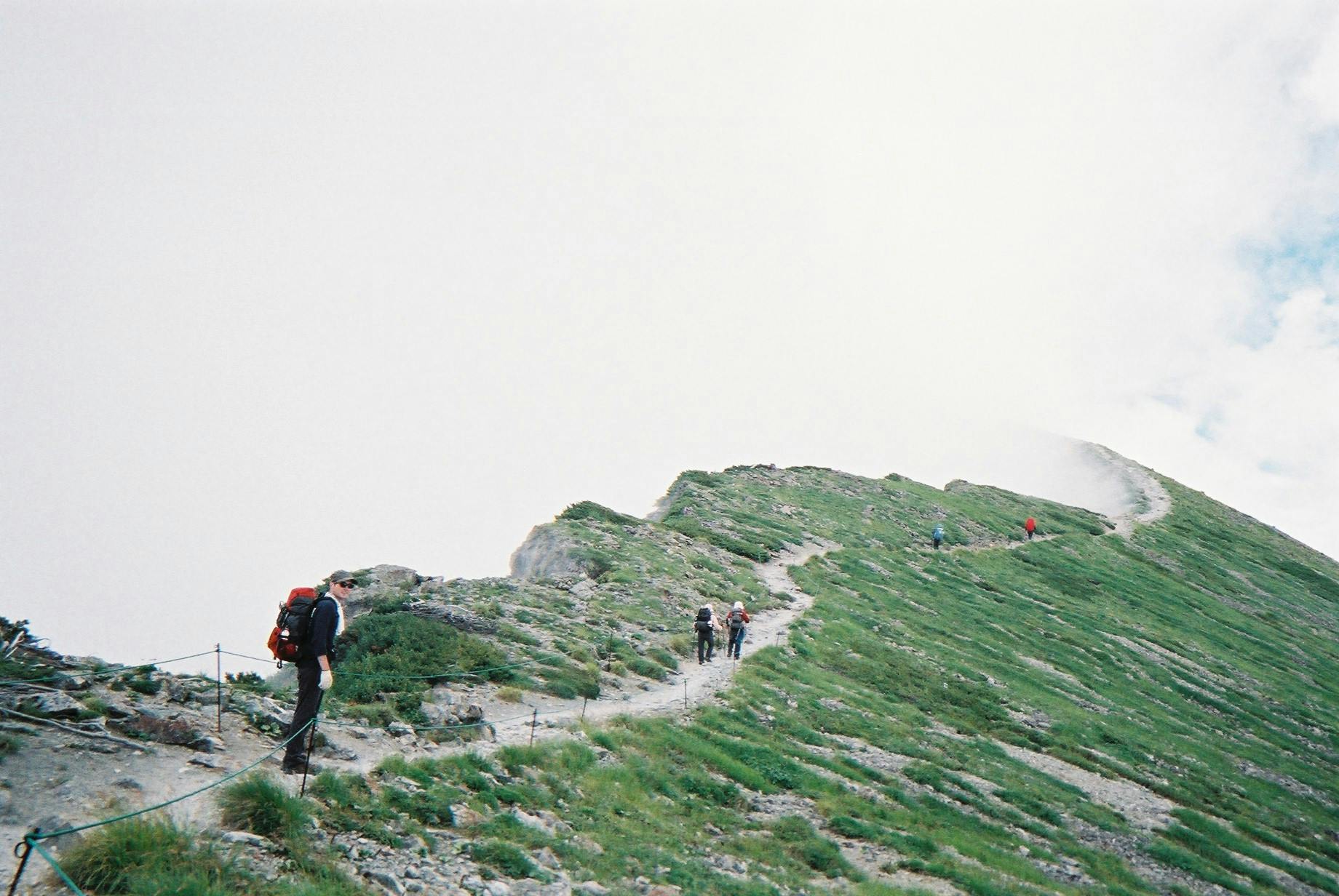 A photo of a few hikers walking along a ridge in the Japan Alps, with clouds spilling over the crest.