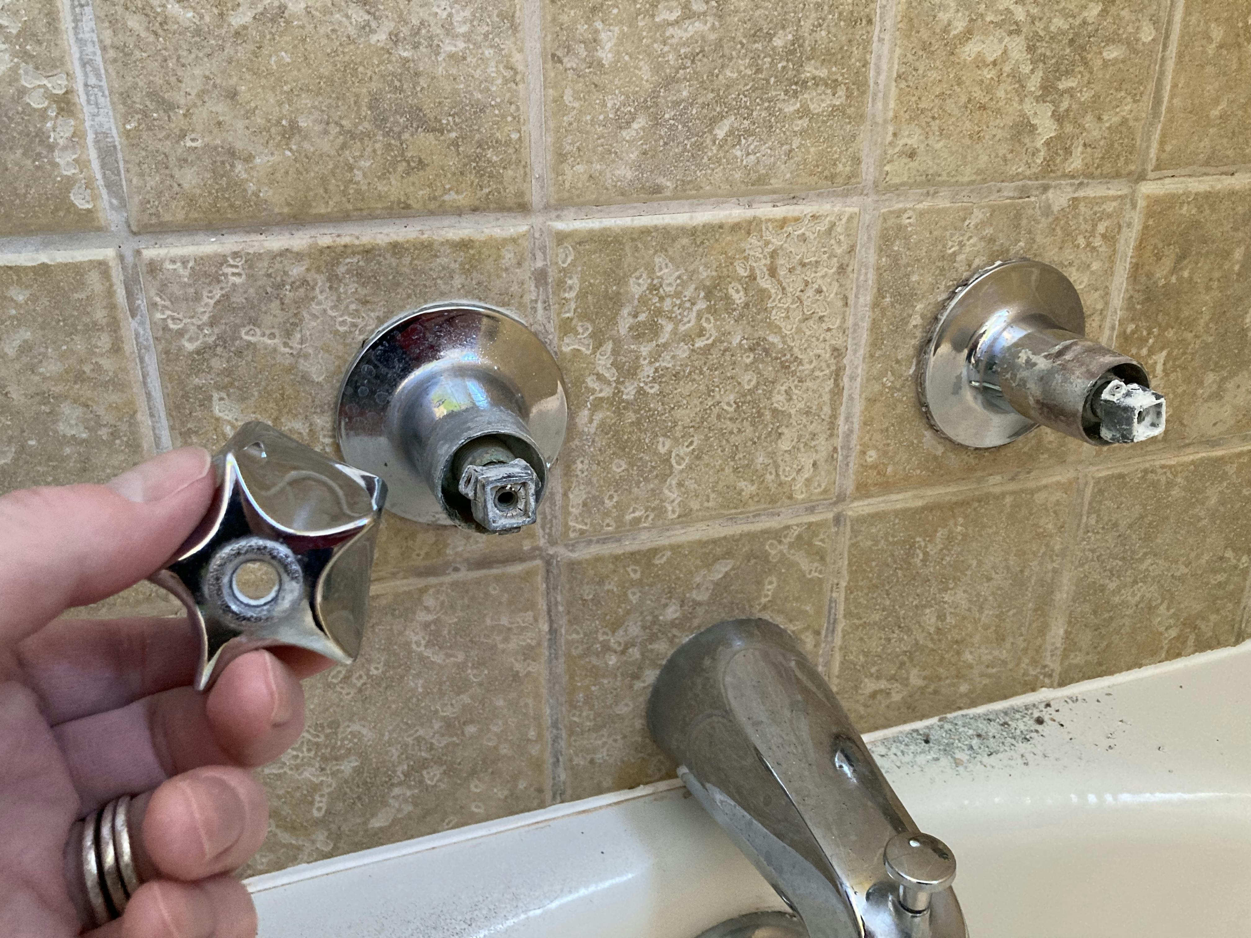 A hand holding the faucet handle of a bathtub faucet in front of a broken bathtub faucet