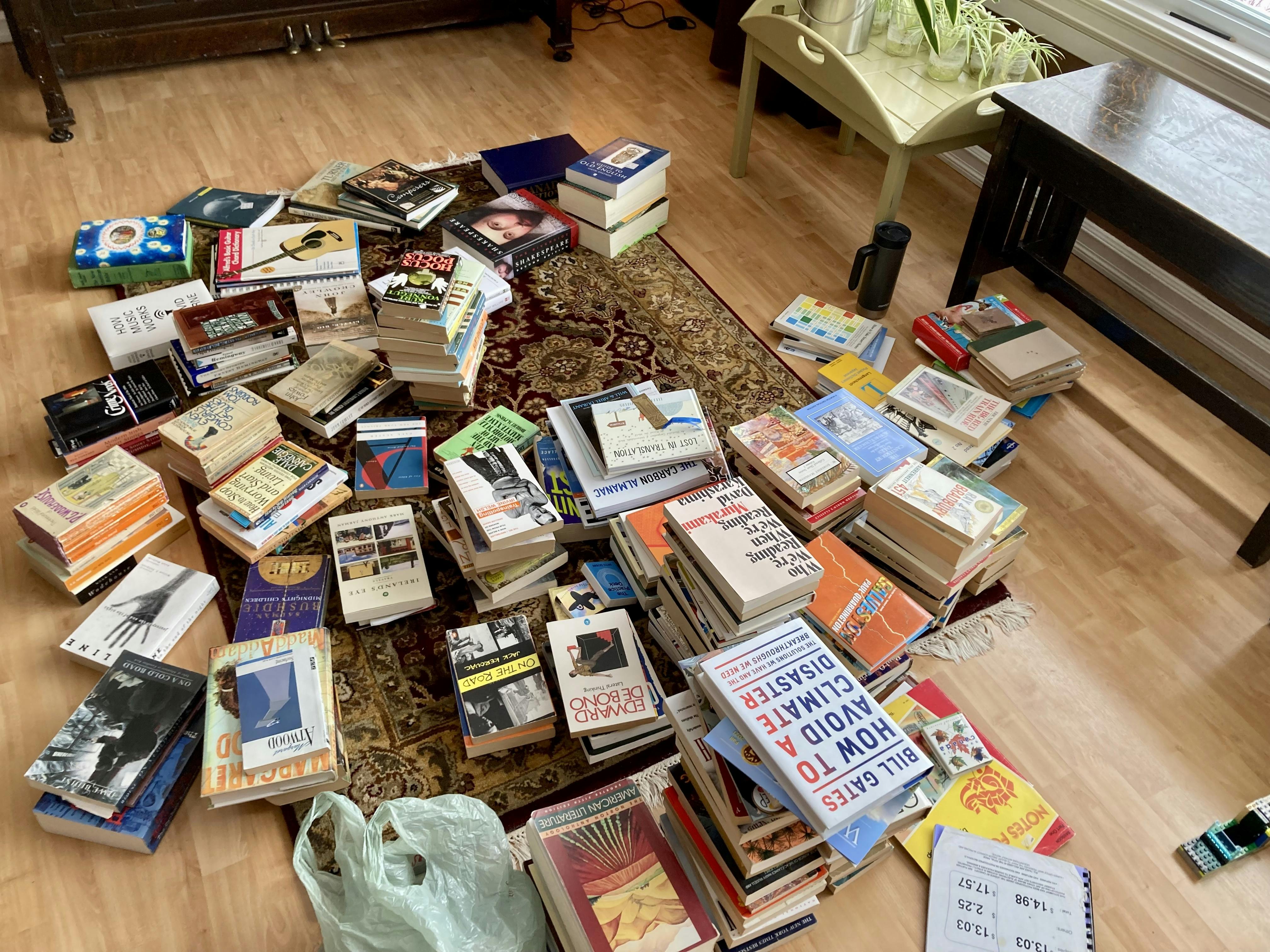 A photo of a whole pile of books spread around a persian rug on a fake hardwood floor.