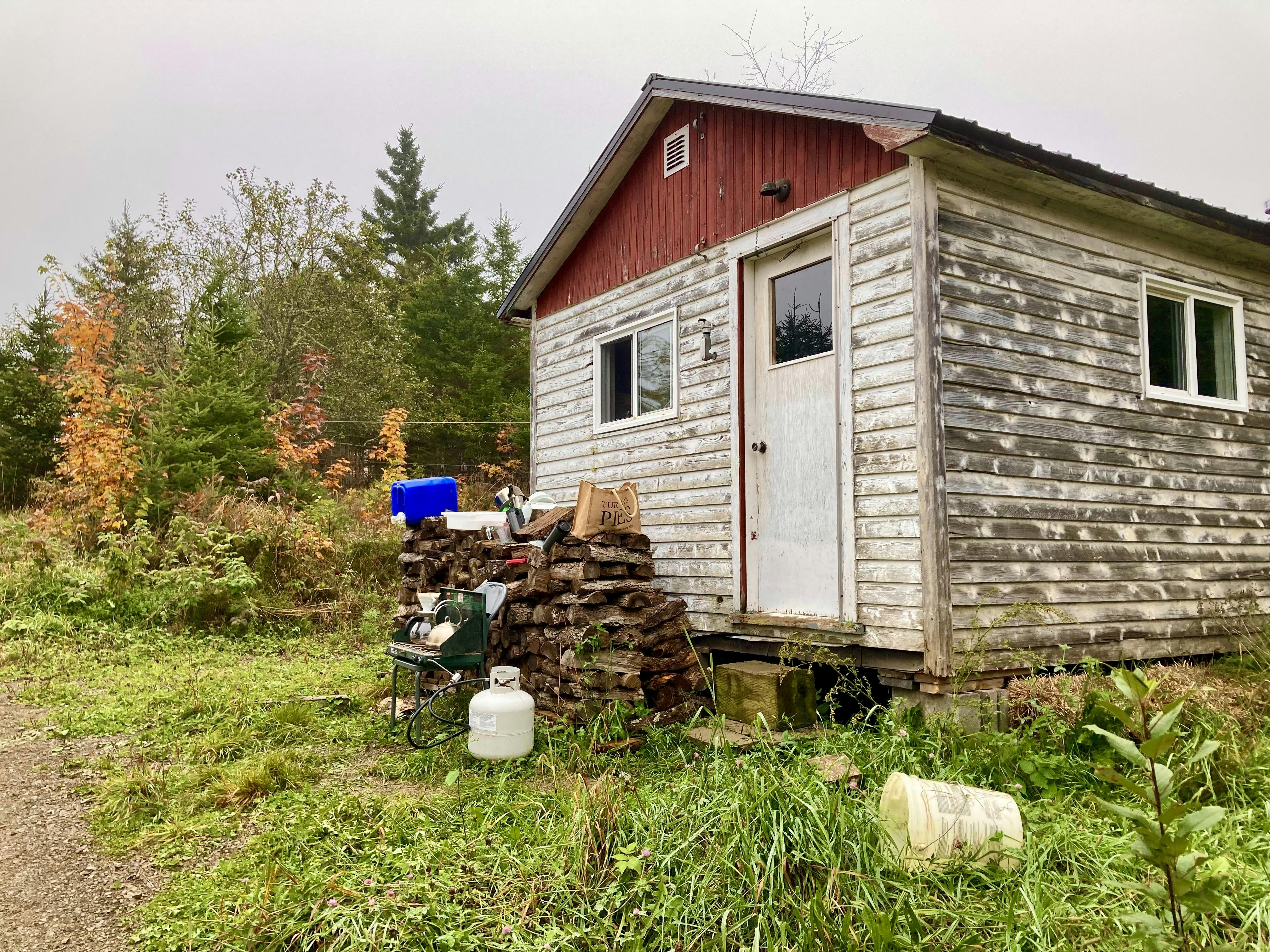 A photo of a small old hunting cabin with a makeshift kitchen set up on a log pile in front of it.