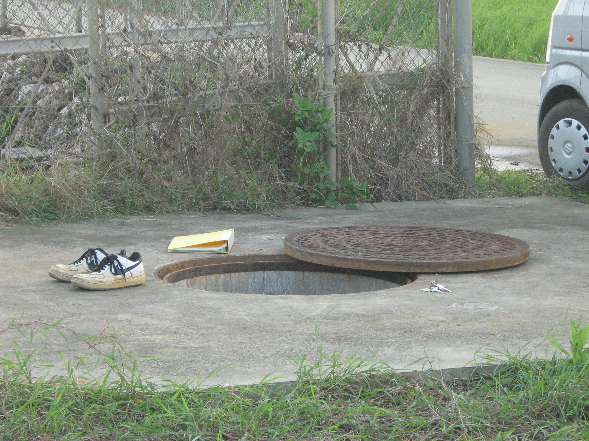 A photo of a sewer entrance hole with the cover removed and a lined up pairg of sneakers, a binder, and a set of keys all sitting around the edge of the hole, with no sign of their owner, who is, presumably, down the hole