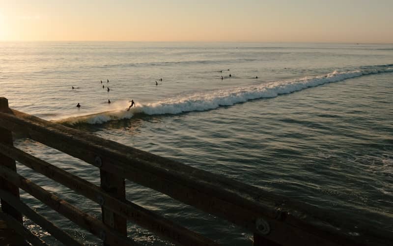 Surfers ride waves near a pier at sunset.