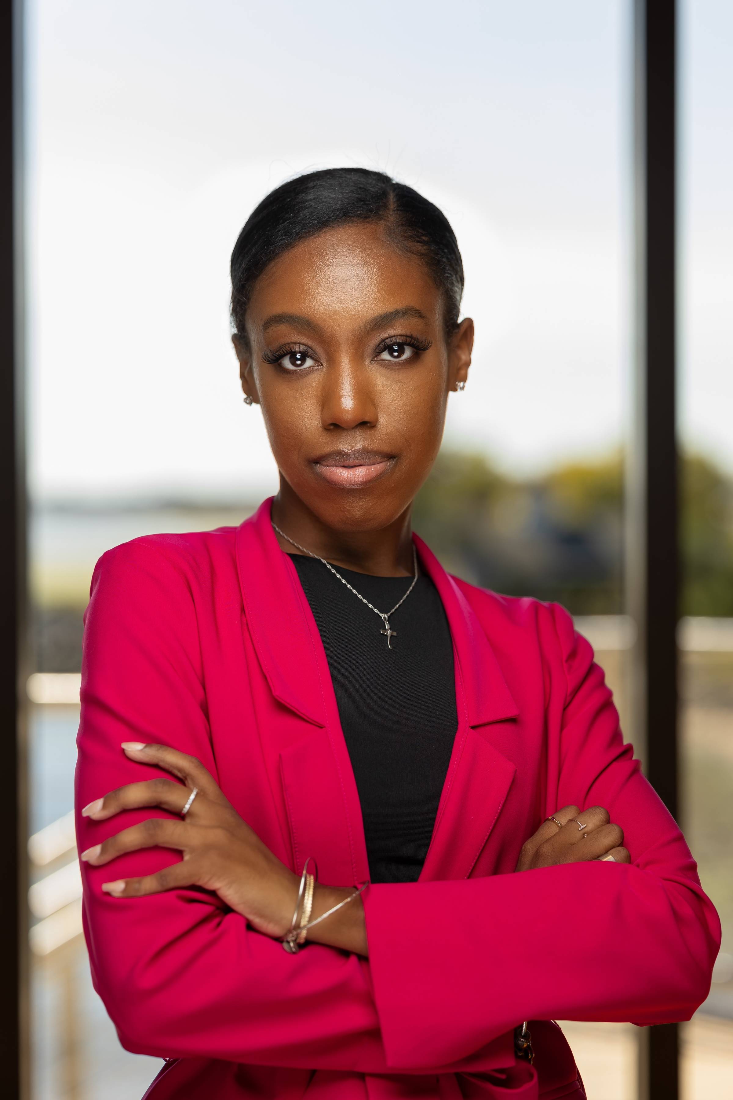 A confident woman stands with her arms crossed, wearing a bright pink blazer over a black top. She has a neat hairstyle and is accessorized with small earrings and a cross necklace. The background is softly blurred, suggesting a window view with natural light.