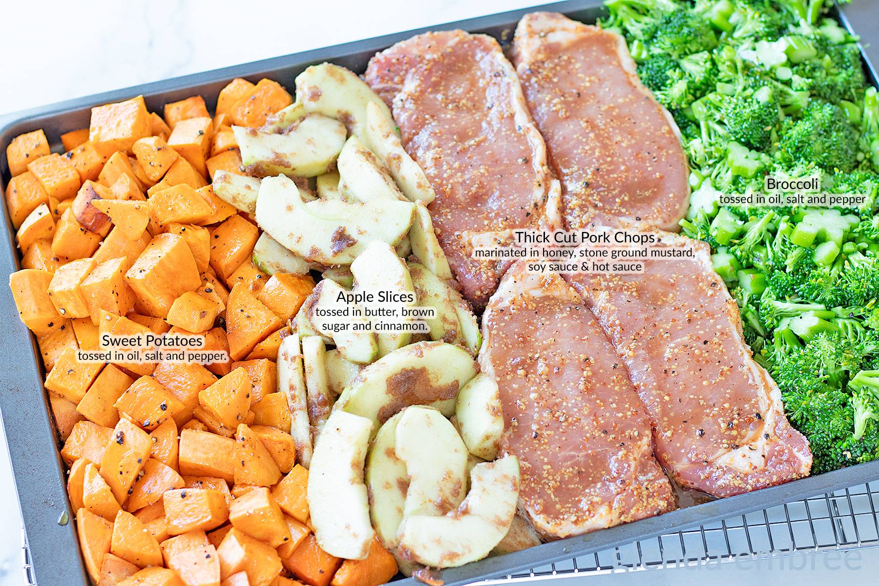 Sheet tray with pork chops, broccoli, sweet potatoes and sliced apples ready to go in the oven to be roasted.
