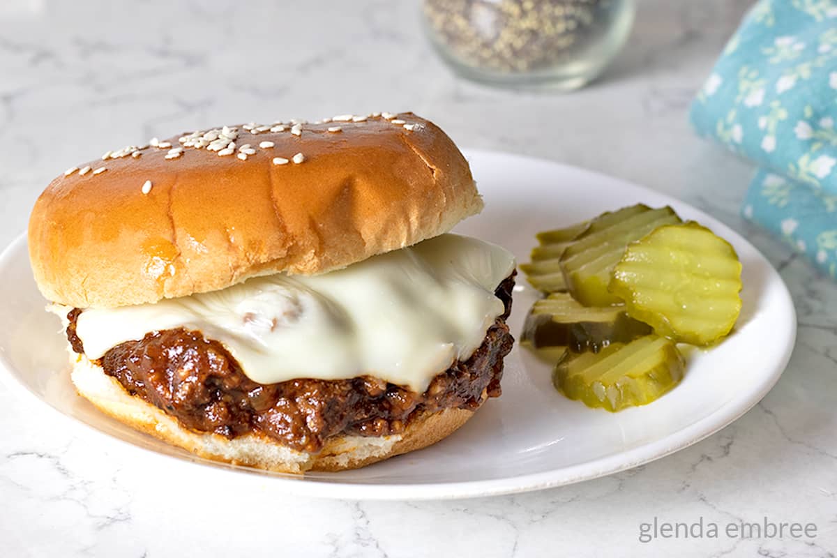 Sloppy Joe sandwich on a white plate with dill pickle chips.