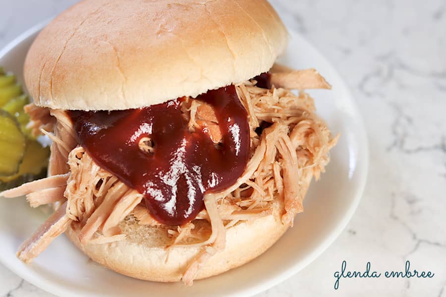 Pulled pork sandwich with BBQ sauce on a white plate with dill pickle chips.