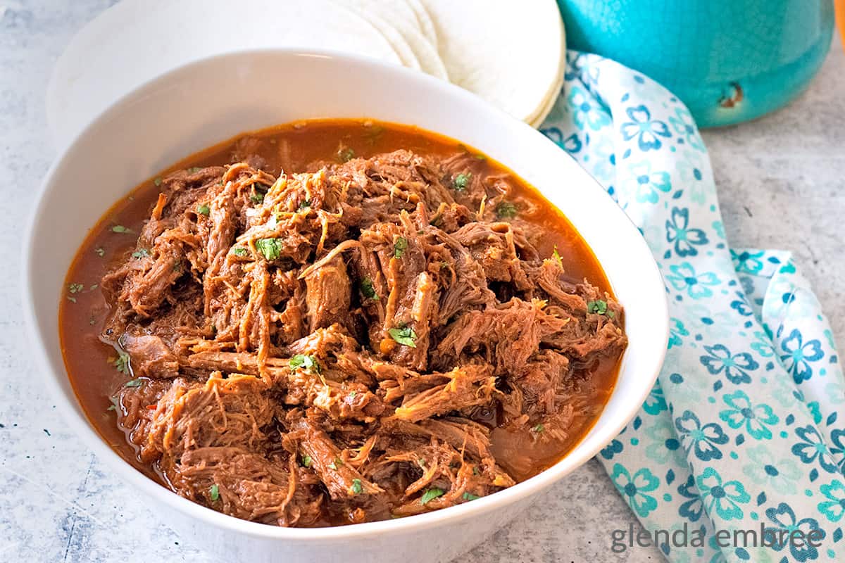 Mexican Shredded Beef in a white ceramic serving bowl next to flour tortillas on a concrete countertop.