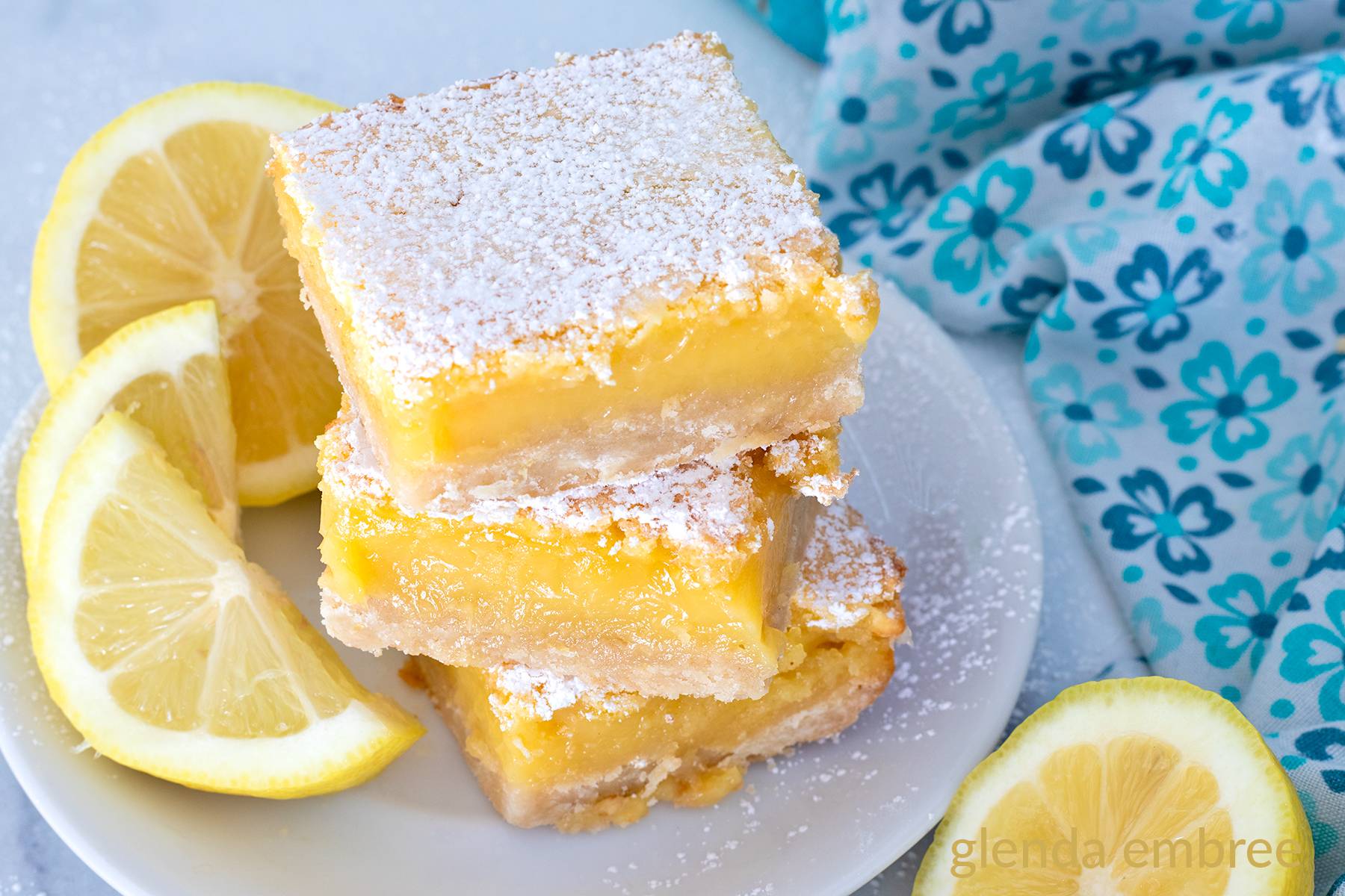 Three lemon bars stacked on a white plate with lemon slices.