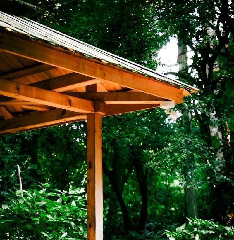 A wooden gazebo structure with a security camera mounted under the roof in a lush forest