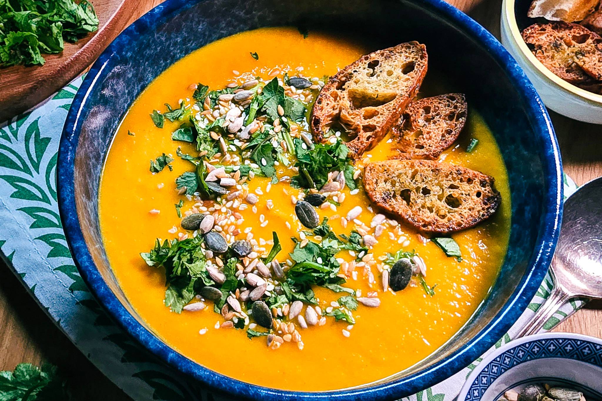 Bright carrot and coriander soup in a blue bowl with mixed seeds, fresh coriander and crisp crostini.