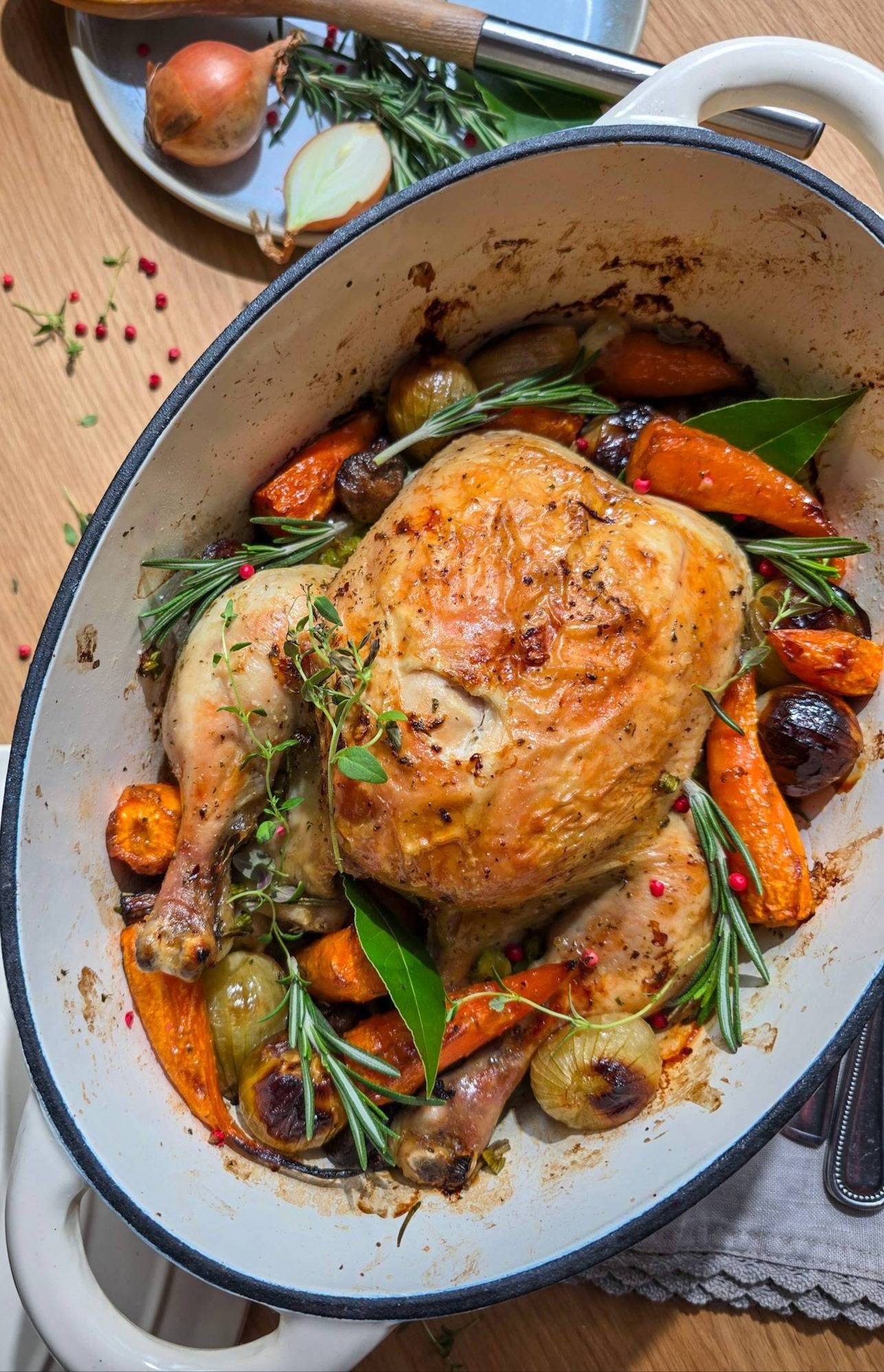 Whole roast chicken in a white cast-iron casserole dish with carrots, onions, rosemary, bay leaves and pink peppercorns on a wooden table, overhead view.